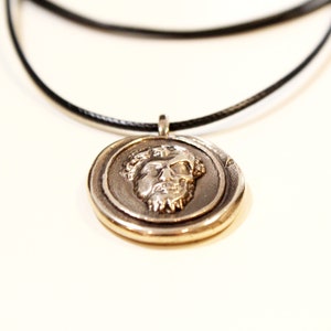 Puede incluir: Un collar con un colgante de plata con un dise&ntilde;o de rostro detallado en un cord&oacute;n negro.