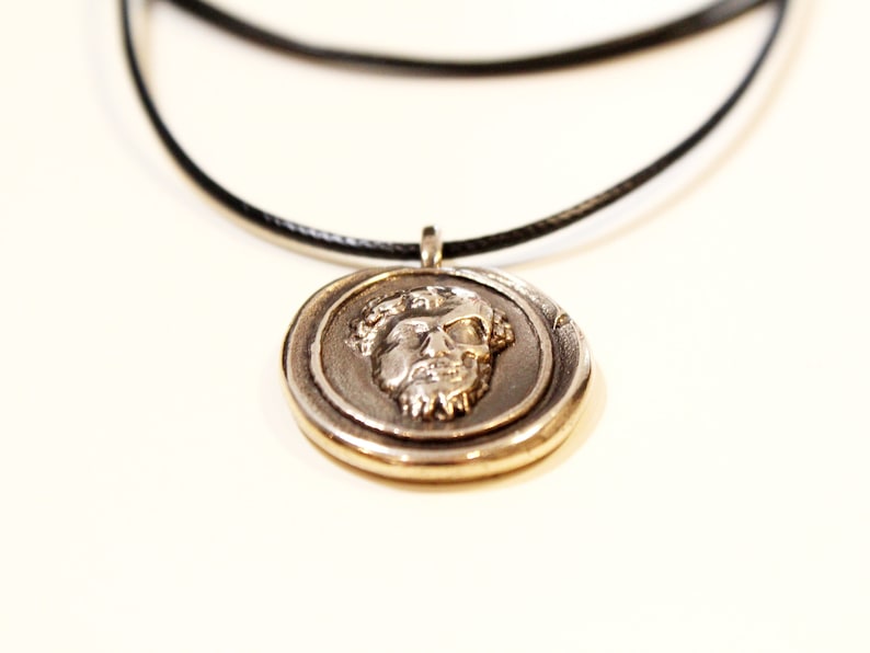 Puede incluir: Un collar con un colgante de plata con un dise&ntilde;o de rostro detallado en un cord&oacute;n negro.