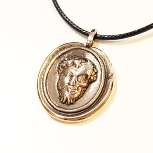 Puede incluir: Un collar con un colgante redondo de color dorado, con un dise&ntilde;o detallado que presenta un rostro esculpido con una corona de hojas. El collar est&aacute; en un cord&oacute;n negro.