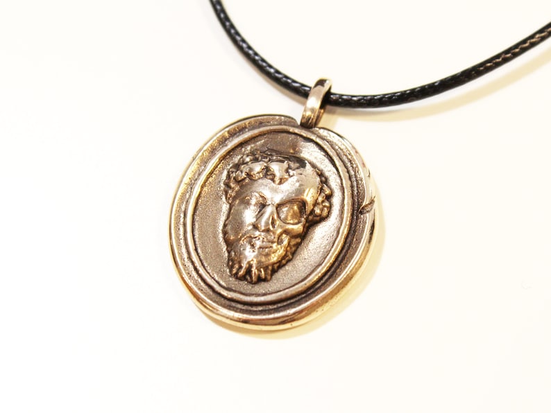 Puede incluir: Un collar con un colgante redondo de color dorado, con un dise&ntilde;o detallado que presenta un rostro esculpido con una corona de hojas. El collar est&aacute; en un cord&oacute;n negro.