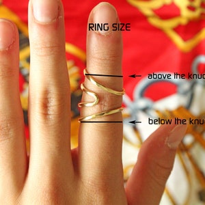 Op de afbeelding: Een hand met een gouden ring met een golvend ontwerp. De ring wordt boven de knokkel gedragen. De tekst "RING SIZE" staat boven de hand. De tekst "above the knuckle" en "below the knuckle" staat aan de rechterkant van de afbeelding.