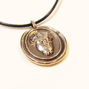 Puede incluir: Un collar con un colgante redondo dorado que presenta un perfil detallado de la cara de un hombre. El colgante est&aacute; en un cord&oacute;n de cuero negro.