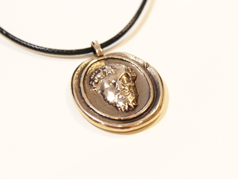 Puede incluir: Un collar con un colgante redondo dorado que presenta un perfil detallado de la cara de un hombre. El colgante est&aacute; en un cord&oacute;n de cuero negro.
