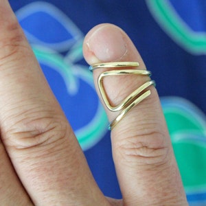 May include: A gold-colored ring with a unique, geometric design. The ring is worn on the index finger of a hand.