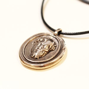 Puede incluir: Un collar con un colgante redondo de plata, con un dise&ntilde;o detallado que presenta un rostro de perfil. El colgante est&aacute; en un cord&oacute;n negro.