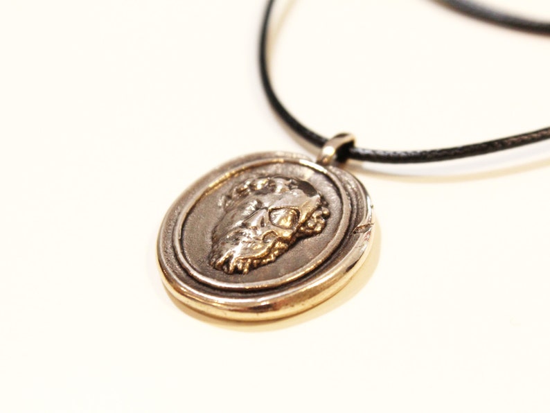 Puede incluir: Un collar con un colgante redondo de plata, con un dise&ntilde;o detallado que presenta un rostro de perfil. El colgante est&aacute; en un cord&oacute;n negro.