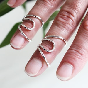 May include: Two silver-toned finger rings with a flowing, abstract design. The rings are worn on the fingers, showcasing their unique, artistic shape. The rings appear to be made of a shiny, metallic material.