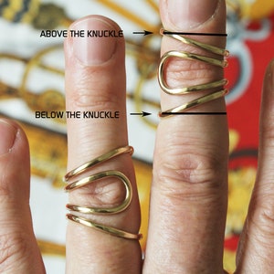 K&ouml;nnte beinhalten: Ein goldfarbener Ring mit einem einzigartigen Design, der sich um den Finger wickelt. Der Ring wird an einem Finger gezeigt, auf dem der Text "ABOVE THE KNUCKLE" und "BELOW THE KNUCKLE" angibt, wo der Ring am Finger sitzen soll.