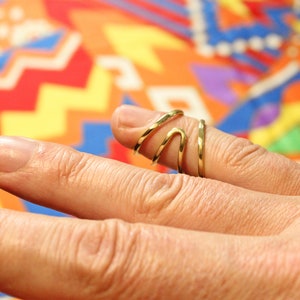 Op de afbeelding: Een gouden ring met een uniek, golvend ontwerp. De ring wordt gedragen aan de middelvinger van een hand.