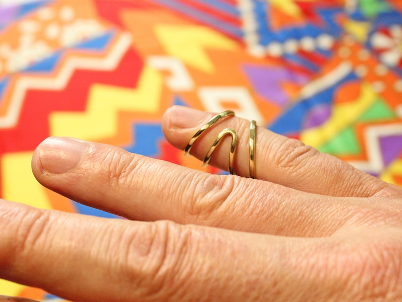 Op de afbeelding: Een gouden ring met een uniek, golvend ontwerp. De ring wordt gedragen aan de middelvinger van een hand.