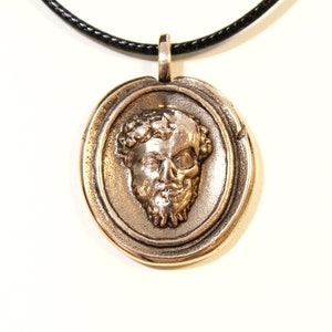 Puede incluir: Un collar con un colgante redondo de plata, con un dise&ntilde;o detallado que presenta una calavera con una corona de laurel. El colgante est&aacute; en un cord&oacute;n negro.