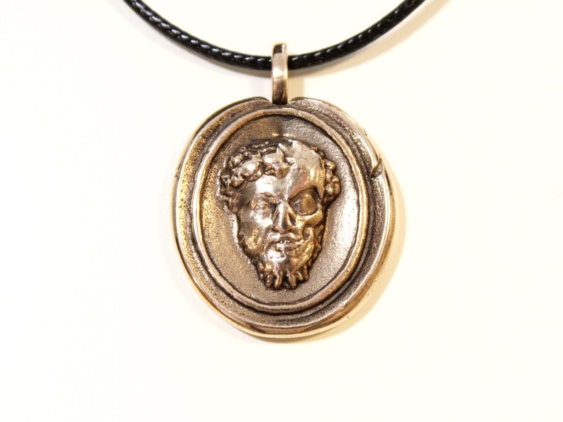 Puede incluir: Un collar con un colgante redondo de plata, con un dise&ntilde;o detallado que presenta una calavera con una corona de laurel. El colgante est&aacute; en un cord&oacute;n negro.
