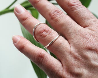 May include: A silver ring is worn on a finger. The ring has a simple, minimalist design, with a curved band that wraps around the finger. The ring is made of a shiny, reflective metal, and it appears to be well-crafted.