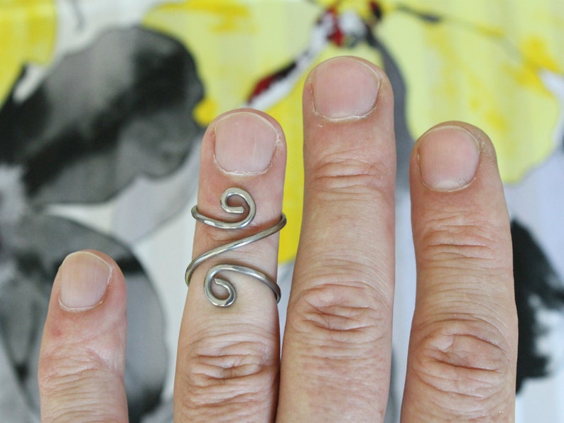 May include: A silver ring with a unique, twisted design. The ring is worn on the middle finger of a hand.