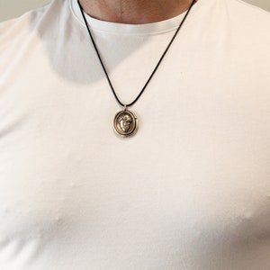 Puede incluir: Un collar con un colgante redondo de color dorado, con un dise&ntilde;o detallado. El colgante presenta una imagen en relieve de una cara con una corona. El collar est&aacute; en un cord&oacute;n negro.