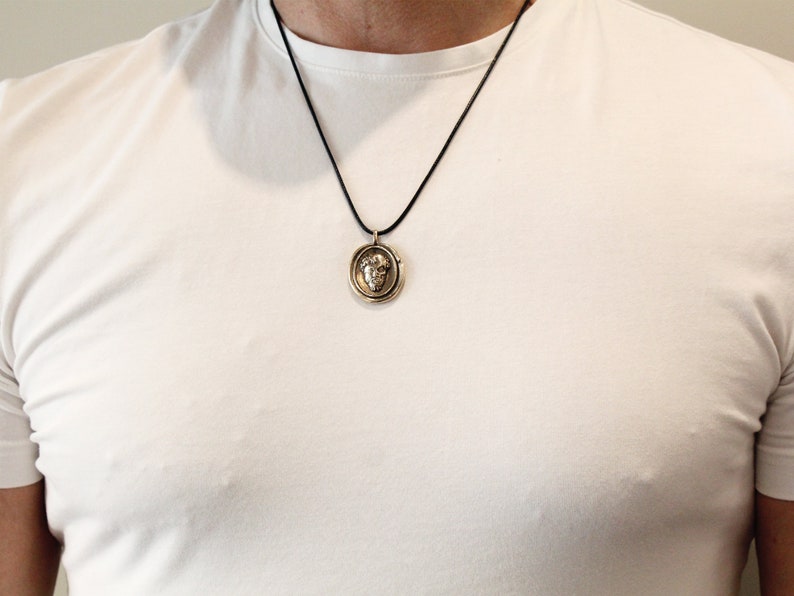 Puede incluir: Un collar con un colgante redondo de color dorado, con un dise&ntilde;o detallado. El colgante presenta una imagen en relieve de una cara con una corona. El collar est&aacute; en un cord&oacute;n negro.
