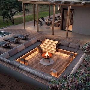 May include: Outdoor living space with a sunken seating area, fire pit, and wooden deck. The seating area has gray cushions and is illuminated by warm lighting. A wooden pergola and surrounding landscape complete the scene.