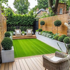 Puede incluir: Un pequeño patio trasero rectangular con una valla de madera, césped verde y una terraza de madera con dos sillas de mimbre y un sillón de mimbre. El patio trasero está rodeado de exuberante vegetación.