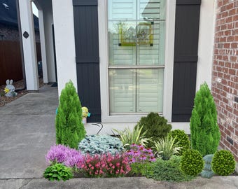 May include: A white house with a black arched window and black shutters. The window has a stained glass design. There is a flower bed in front of the house with pink and white flowers, green bushes, and a small pine tree.