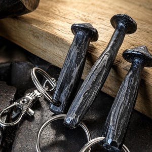 May include: Three handcrafted, dark metal keyrings with textured, nail-shaped pendants. Each keyring has a silver ring and a clasp. The keyrings rest on a wooden surface, with a hammer visible in the background.