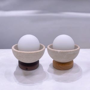 Könnte beinhalten: Zwei weiße Keramiklampen mit matten Glaskugeln. Die Lampen haben eine beige und braune gesprenkelte Oberfläche und stehen auf Holzsockeln.
