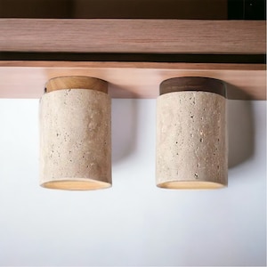 May include: Two beige cylindrical light fixtures with a wood top and bottom, mounted to a wooden shelf.