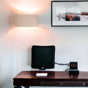 May include: A dark brown wooden desk with a black cushion attached to the wall, a black electrical outlet with a cord running to the cushion, and a white rectangular object on the desk. A white ceiling light fixture is mounted on the wall above the desk.