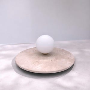Pode incluir: Uma luminária de globo branco fica sobre uma base redonda de travertino bege. A luminária é feita de vidro e tem uma base de latão.