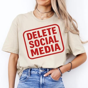Peut inclure: T-shirt beige à manches courtes avec un motif carré rouge portant l'inscription blanche en gras "DELETE SOCIAL MEDIA". Le t-shirt est en matière souple et convient aux tenues décontractées. Le modèle porte un jean bleu.