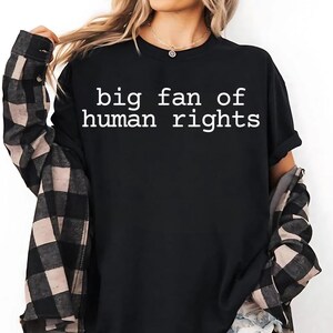 Może przedstawiać: Czarny t-shirt z białym napisem "big fan of human rights". Koszulka jest połączona z czarno-beżową koszulą w kratę zapinaną na guziki. Modelka ma na sobie złoty naszyjnik.