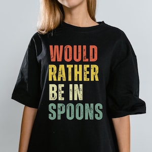 Puede incluir: Camiseta negra con la frase "WOULD RATHER BE IN SPOONS" en una fuente retro. El texto es de color naranja, amarillo y verde. La camiseta tiene mangas cortas y cuello redondo.