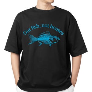 Op de afbeelding: Zwart T-shirt met een blauwe afbeelding van een vis en de tekst "Gut fish, not houses". Het ontwerp bevat een gedetailleerde illustratie van een vissenskelet. Het shirt heeft korte mouwen en een ronde hals.