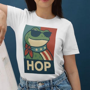Puede incluir: Camiseta blanca con una rana de dibujos animados que lleva gafas de sol, una pajarita roja y una bufanda con estrellas y rayas. La palabra "HOP" está impresa en letras negritas debajo de la ilustración de la rana. La camiseta es de material suave.