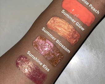 May include: Five different lip glosses swatched on a person's arm. The glosses are labeled "Sunshine Kiss", "Summer Blossom", "Summer Glow", and "Paradise Peach".