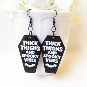 May include: Black coffin-shaped earrings with the text "THICK THIGHS AND SPOOKY VIBES" in white. A white bat graphic is below the text. The earrings hang from black metal hooks and are displayed against a white background.