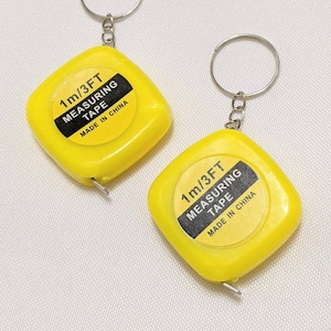 Puede incluir: Dos llaveros con cinta métrica amarillos. Cada llavero tiene una caja amarilla de forma cuadrada con una etiqueta en blanco y negro que dice "1m/3FT MEASURING TAPE MADE IN CHINA". La cinta mide 1 metro.