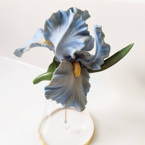 May include: A blue porcelain iris flower figurine with green leaves sits atop a clear glass bell with a gold rim. A crystal hangs from a chain attached to the bell.