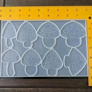 May include: A clear silicone mold featuring eight mushroom-shaped cavities. The mold includes a ruler for size reference. Each mushroom design has a different pattern of circular details on the cap. The mold is designed for crafting.