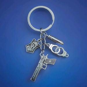 May include: Silver-toned keychain with a large ring and several charms. Charms include a police badge, a handgun, a bullet, and handcuffs. The word "Freedom" is visible on the handcuffs. The background is a solid blue.