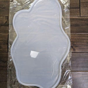 Może przedstawiać: Przezroczysta, silikonowa forma w kształcie chmury w przezroczystej plastikowej torbie. Forma ma gładką powierzchnię i zaokrąglone krawędzie. Tło to drewniana powierzchnia.