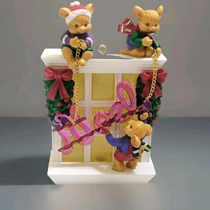 Puede incluir: Una figura decorativa con temática navideña que presenta tres figuras de ratones. Los ratones visten ropa de invierno y sostienen cadenas que suspenden la palabra "love". La figura tiene un diseño de ventana con coronas y lazos.