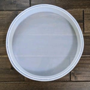 May include: A round, white plastic tray with a mesh center. The tray has a raised edge and is designed for straining or sifting. The tray is shown on a wooden surface.