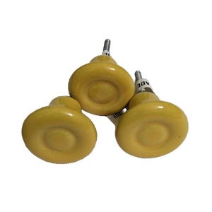 Könnte beinhalten: Drei gelbe Keramik-Schrankknöpfe mit einer silbernen Schraube. Die Knöpfe haben ein erhabenes, kreisförmiges Design.