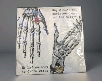 May include: A package of white paper napkins with a skeleton hand design. One hand wears a ring with a pink gem, and the other wears a watch. Text reads, "Why didn't the skeleton dance at the party? He had no body to dance with!"