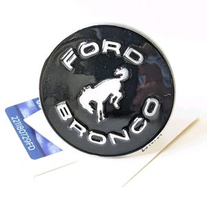 May include: A black and silver Ford Bronco emblem. The emblem is circular with the word "FORD" at the top and "BRONCO" at the bottom in silver lettering. A white horse graphic is in the center.