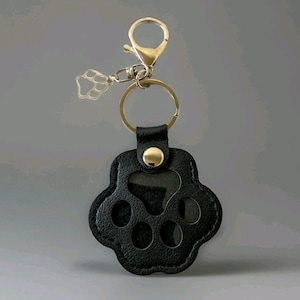 Op de afbeelding: Zwarte leren sleutelhanger met pootafdruk en goudkleurige hardware. De sleutelhanger heeft een gouden karabijnhaak, een kleine pootafdrukbedel en een sleutelring. Het pootafdrukontwerp heeft uitgesneden details.