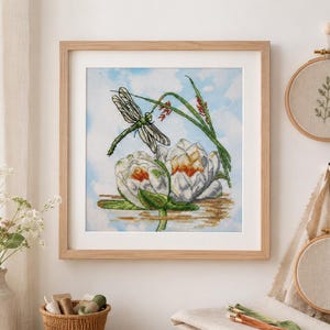 May include: A framed cross-stitch of a dragonfly and water lilies. The dragonfly is green and gray, perched above two white and yellow lilies. The background is a light blue sky. The frame is light wood.