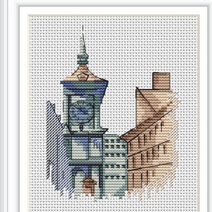 May include: Cross-stitch artwork of a European cityscape, featuring a clock tower and buildings in shades of blue, green, and brown. The piece is framed in a white border, showcasing the detailed needlework and architectural elements.