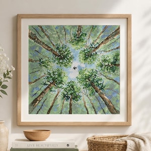 May include: A framed artwork featuring a view looking up through a forest canopy. The artwork depicts tall trees with green foliage and brown trunks, with a bird in the center against a blue sky. The frame is made of light-colored wood.