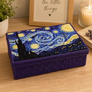 May include: A rectangular, purple box with a cross-stitch design depicting Van Gogh's "Starry Night." The lid features the swirling blue and yellow night sky, with a dark silhouette of a town. The box is on a wooden surface.